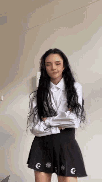 a woman with long black hair wearing a white shirt and black skirt is standing with her arms crossed .
