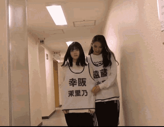 two girls are walking down a hallway with one wearing a white shirt that says ' lucky ' on it