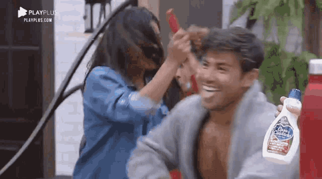 a man is holding a bottle of syrup while a woman brushes his hair .