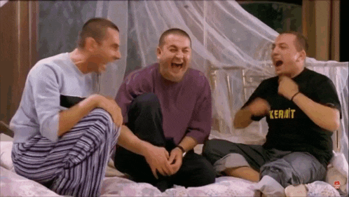 three men are sitting on a bed laughing and one has a shirt that says kermit