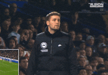 a man wearing an american express jacket stands in front of a soccer field