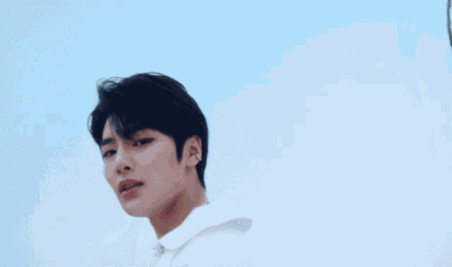 a young man wearing a white shirt and a necklace looks at the camera with a tree in the background