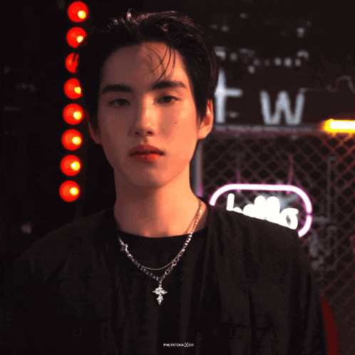 a young man wearing a black shirt and a cross necklace is standing in front of a neon sign that says t w