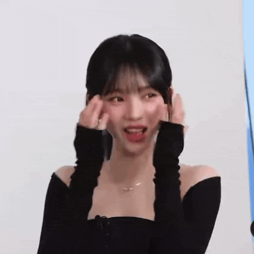 a woman in a black off the shoulder top is smiling and covering her ears with her hands .