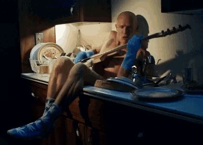 a shirtless man is playing a guitar in a messy kitchen
