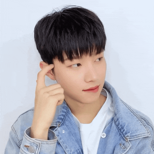 a young man wearing a blue denim jacket and a white shirt holds his hand to his ear