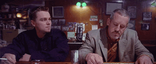 two men are sitting at a bar with one smoking a cigarette and the other drinking a drink
