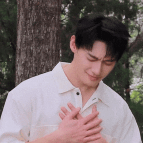 a man in a white shirt is holding his hands to his chest in front of a tree .