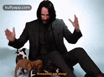 a man in a suit is sitting on the floor with a bunch of puppies and saying `` everybody get along ! ''