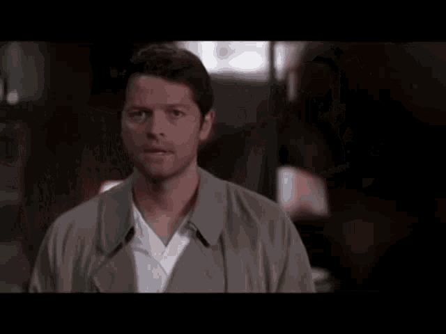 a man in a trench coat and white shirt is standing in a dark room looking at the camera .