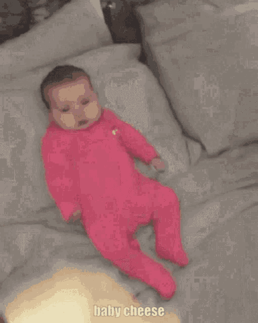 a baby in a pink outfit is laying on a bed with the words baby cheese written on the bottom .