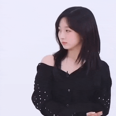 a young woman wearing a black off the shoulder sweater with pearls on the sleeves is looking at the camera .