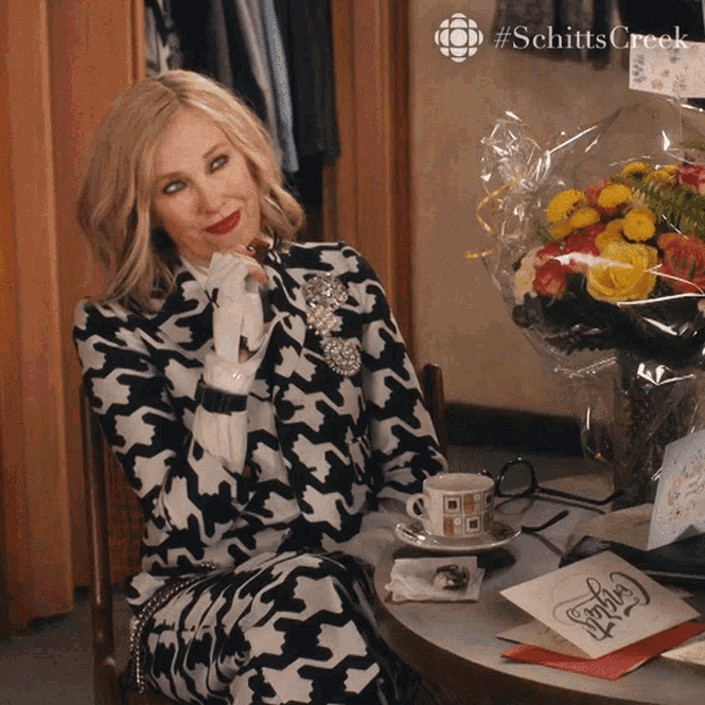 a woman sits at a table with a bouquet of flowers and a card that says schitts creek on it