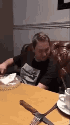 a man in a black shirt is sitting at a table with a knife and fork .