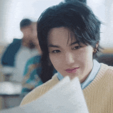 a young man in a yellow sweater is reading a piece of paper in a classroom .