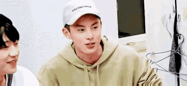 a man wearing a balenciaga hat and a hoodie is sitting next to another man .