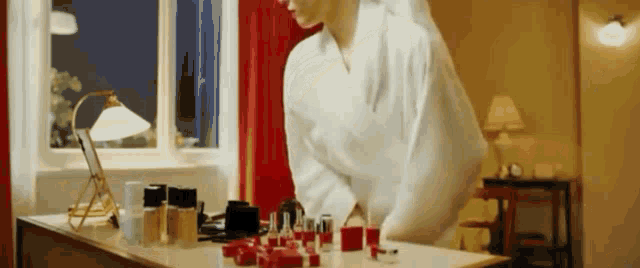 a woman in a bathrobe is looking at herself in a mirror while sitting at a table with cosmetics on it .