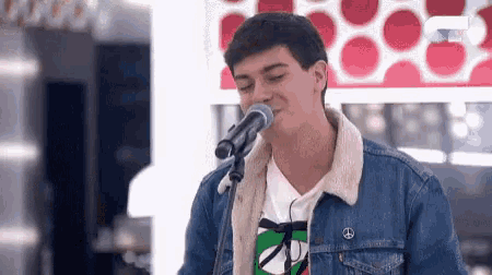 a young man is singing into a microphone while wearing a denim jacket and a peace sign shirt .