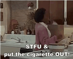 a woman in a pink sweater is standing in a kitchen with the words stfu and put the cigarette out .