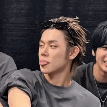 a man wearing sunglasses sticks out his tongue while another man looks on