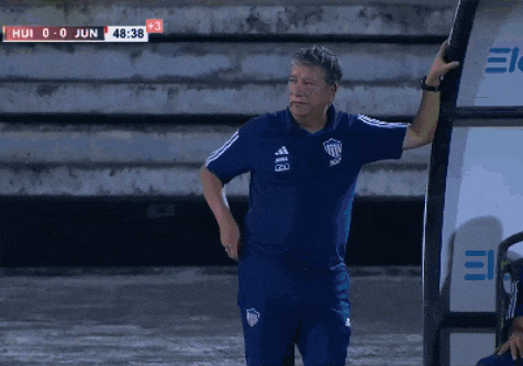 a man wearing a blue adidas shirt stands in front of a scoreboard that says hui 0-0 jun