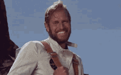 a man with a beard and suspenders is smiling while standing in front of a mountain .