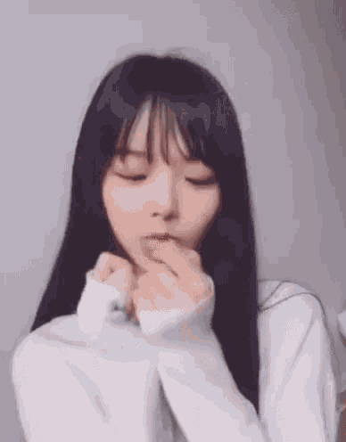 a girl with long black hair and bangs is wearing a white sweater and covering her mouth with her hands .