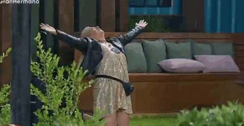 a woman in a gold dress and a leather jacket is standing in front of a wooden bench with her arms outstretched .