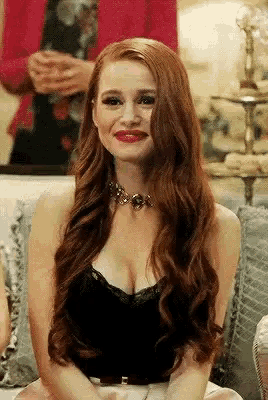 a woman with long red hair is wearing a black top and a necklace while sitting on a couch .