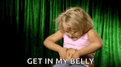 a little girl is holding her belly in front of a green curtain and says `` get in my belly '' .