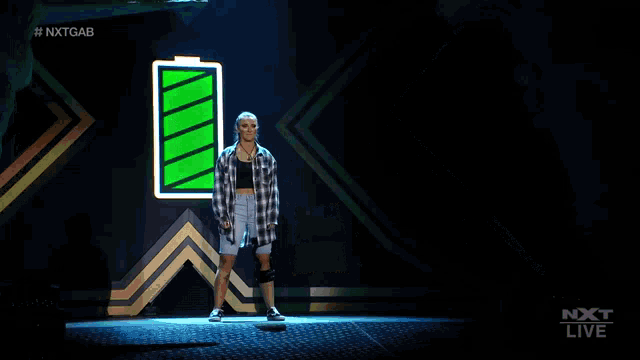 a woman in a plaid shirt and shorts stands on a stage with her arms outstretched in front of a green battery .