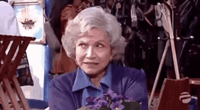 an elderly woman is sitting at a table with purple flowers and a laptop .