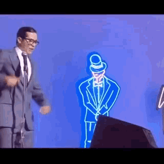a man in a suit and tie is standing in front of a microphone with a neon man behind him