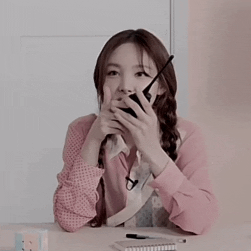 a woman in a pink shirt is holding a cell phone to her mouth