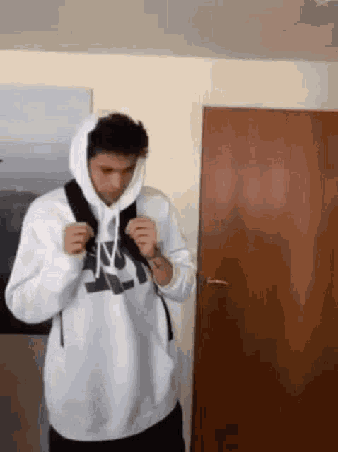 a man in a white hoodie is standing in front of a door holding a backpack .