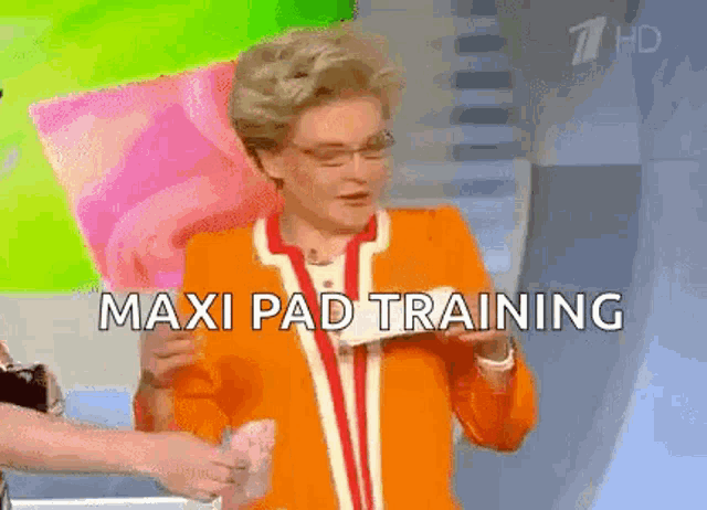 a woman in an orange jacket is holding a pink pillow and the words maxi pad training are above her .