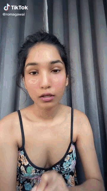 a woman in a floral tank top is standing in front of a grey curtain and looking at the camera .