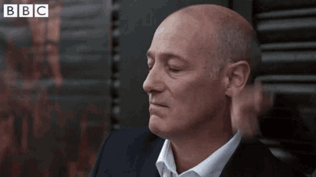 a bald man in a suit and white shirt is sitting in front of a window with his eyes closed .