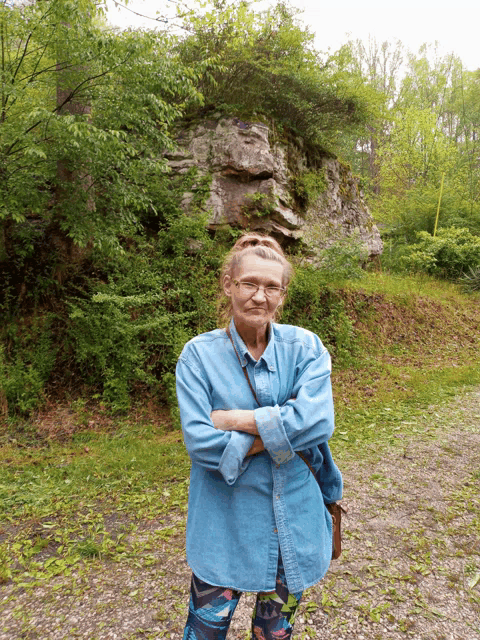 a woman in a blue shirt stands with her arms crossed in front of a large rock