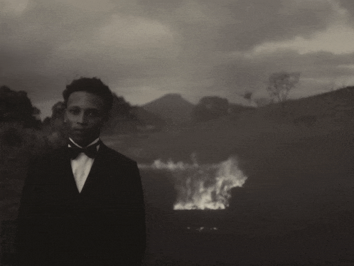 a man in a tuxedo stands in front of a burning field