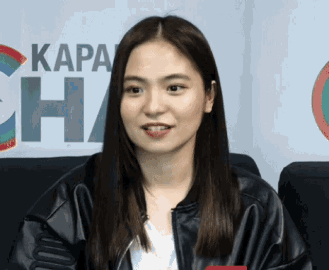 a woman wearing a black leather jacket is smiling in front of a sign that says kapa