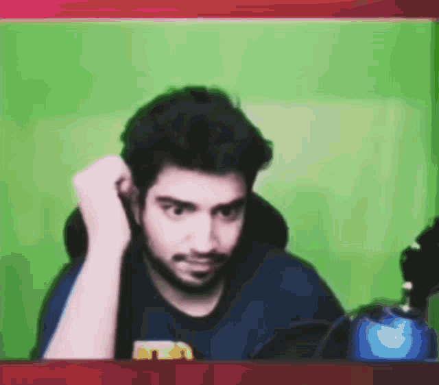 a man with a beard is sitting in front of a green screen with his hand on his ear .