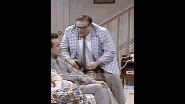 a man in a plaid jacket and tie is talking to another man in a chair