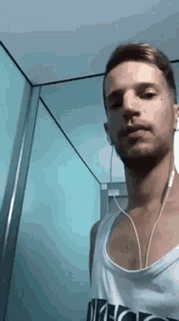a man wearing a white tank top and ear buds is standing in a bathroom .