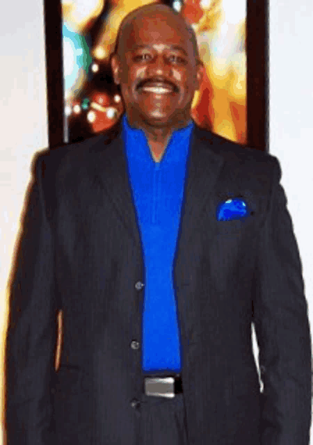 a man in a suit and blue shirt smiles in front of a picture