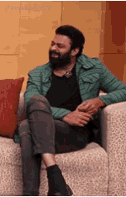 a man with a beard is sitting on a couch wearing a green jacket .