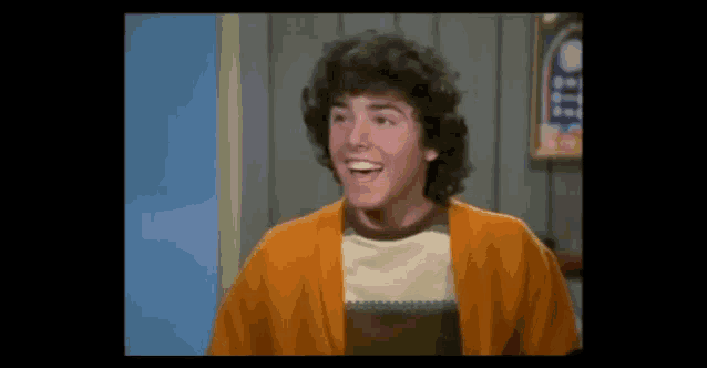 a young man with curly hair is wearing a yellow cardigan and smiling .