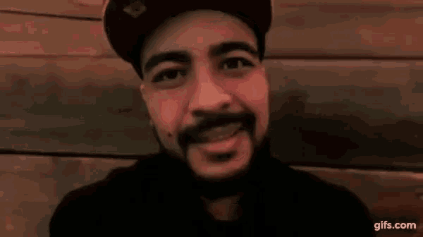 a man with a beard is wearing a hat and giving a thumbs up sign .