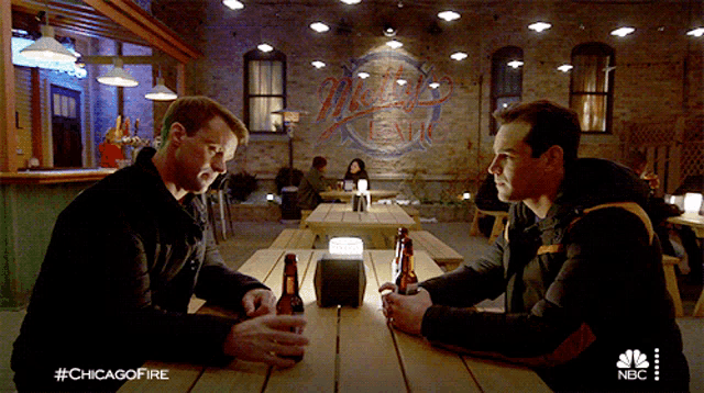 two men sit at a picnic table with bottles of beer in front of a mural that says mets