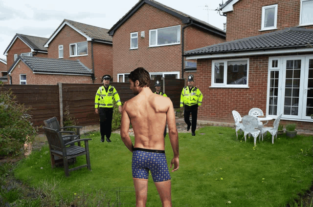 a shirtless man in boxer shorts is standing in a backyard with two police officers